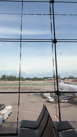 The video shows a view from inside an airport terminal through a large glass window. Outside, an airplane is seen taking off and ascending into the sky. The airport tarmac and some parked vehicles are visible in the foreground, with trees and a glimpse of the ocean in the distance. The sky is partly cloudy.