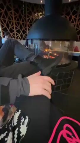 The video shows a person sitting with their legs crossed and their left foot resting on the edge of a modern, circular fireplace. The person is wearing black pants and a black boot on the left foot. The fireplace has a black dome-shaped hood and a glass enclosure around the fire. Visible flames are inside the fireplace. The person's hands are resting on their right leg. Part of a black and white patterned garment is visible on the left. A bright pink cord is also visible. In the background, other people are sitting on a couch. The room has a decorative, geometric pattern on the wall.