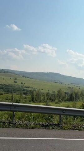 A wide shot captures a lush green landscape with rolling hills under a clear blue sky dotted with white clouds. A silver guardrail runs horizontally across the foreground, separating a paved road from the grassy verge. The camera pans slowly to the right, revealing more of the verdant scenery, including scattered trees and a dense forest in the mid-ground. The road appears to be a highway or main route, indicated by the guardrail and asphalt surface. The overall impression is one of a serene, natural environment on a bright, sunny day.