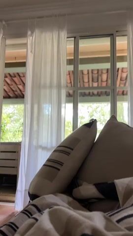 The camera pans across the interior of a room. A couch with pillows and a blanket is in the foreground. There's a large wooden coffee table with a lamp, laptop, vase and teacup. Beyond the table, a blonde woman is sitting in a chair. Large sliding glass doors with sheer white curtains lead to a patio with a wood floor and a red tile roof. There are trees visible outside. The room has terracotta tile floors. A framed picture hangs on the wall.