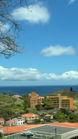 The video shows a sunny, bright day with a view overlooking a coastal landscape. The sky is blue with scattered white clouds. A large body of water, presumably the ocean, is visible in the background with a hint of land in the far distance. In the middle ground, there are several buildings, primarily in shades of orange and with red-tiled roofs, nestled amongst green vegetation. A road winds through the landscape. In the foreground, there are rooftops of buildings, with some vegetation and bare tree branches visible on the left side of the frame. The overall scene suggests a peaceful and scenic location.