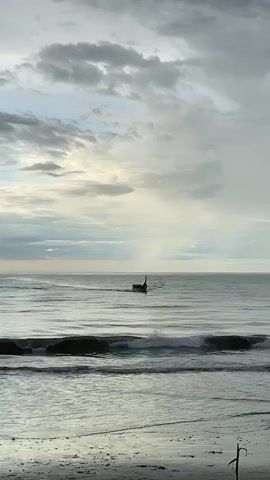 A lone boat sails on the ocean under a cloudy sky. The waves are gently rolling towards a sandy beach.