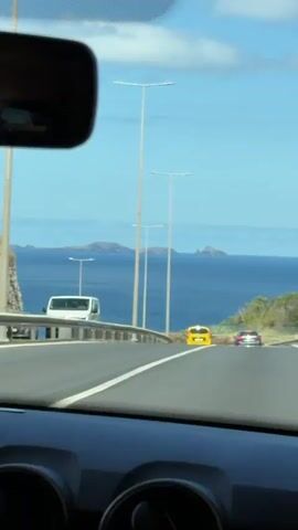The video shows a view from the inside of a car driving on a highway. The camera is focused through the windshield, and the dashboard is visible. The car is driving alongside the ocean. In the distance, there are a few islands visible on the horizon. There are some light posts alongside the road. There is a car ahead. The sky is bright blue with a few clouds. The rear-view mirror is visible in the top left.