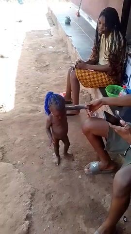 A video shows a small child standing on dirt in front of a cement step. The child is Black and has blue braids. The child is nude. Three young adults sit on the cement step. One is female, with long hair, wearing a brown shirt and a yellow skirt with a brown pattern. Two are male. One male is shirtless and is holding a smartphone. Both men wear shorts and sandals. The shirtless man is smiling. The child extends an arm toward the shirtless man. The woman on the cement step and the other man are also smiling. A green bowl, a red bottle, and a white box are on the cement step behind the people.