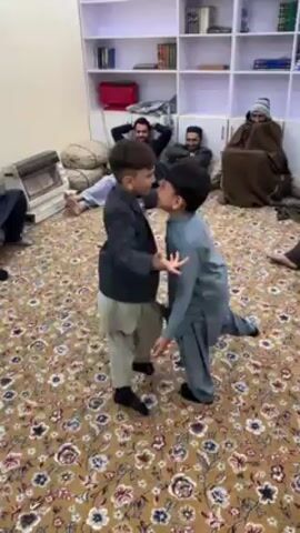 In a carpeted room, two young boys are dancing, facing each other and moving their arms. A group of men sit around the perimeter of the room, observing. One man is reclining and smiling. The room is furnished with a white bookshelf filled with books and other items. There's also a small heater and stacked cushions in the background. The carpet has a complex, ornate pattern.