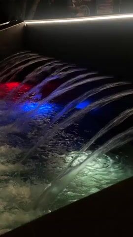 A water fountain with colored lights at night. The fountain has multiple streams of water cascading down into a pool. The lights are red, blue, and white, illuminating the water and creating reflections. In the background, there are silhouettes of people sitting and standing, and trees. The overall atmosphere is dark and atmospheric.