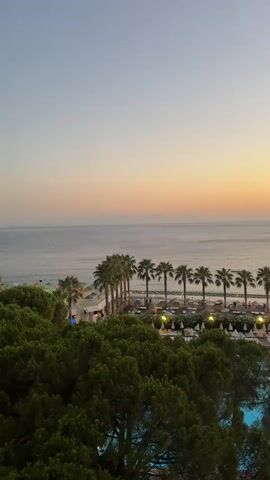 The video shows a scenic view of a beach resort at sunset. The foreground is dominated by lush green trees, with a glimpse of a blue swimming pool to the right. Beyond the trees, a row of palm trees lines a beach area. The beach has numerous lounge chairs and umbrellas set up, with some people visible walking around. The sea stretches out to the horizon, where the soft hues of orange and pink from the setting sun create a beautiful gradient in the sky. Distant mountains are faintly visible across the water. The overall atmosphere is peaceful and relaxing, capturing the tranquility of a tropical evening.