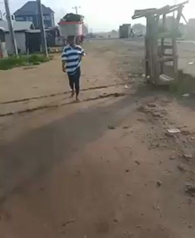 The video shows a person walking down a dirt road. The person is wearing a blue and white striped shirt, black pants, and sandals, and is carrying a large, clear container on their head. The road is dirt, with several tracks running parallel to each other along its length. There are buildings and structures visible in the background, including a corrugated metal structure on the left side of the screen, and a more substantial building with a grey roof further in the distance. There's also what looks like a wooden frame or pallet sitting on the right side of the road. The overall impression is of a basic, possibly rural or developing area. The sky appears overcast, and the lighting suggests it is either early morning or late afternoon.