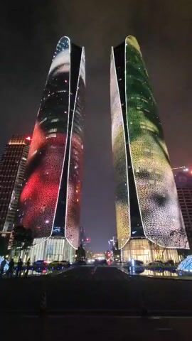 The video shows two tall, modern skyscrapers at night. Both buildings are illuminated with colorful LED lights that create dynamic patterns across their facades. The left building displays a swirling pattern of red, pink, and white lights, resembling a nebula or a galaxy. The right building features a pattern of green, yellow, and white lights, possibly depicting foliage or a starry sky. The buildings are positioned side-by-side, facing a street with cars parked and driving by. The streetlights and car headlights are visible, casting reflections on the wet pavement. There are a few people standing on the sidewalk in front of the buildings, observing the light show. The overall scene is a vibrant and modern urban landscape with a striking architectural and light display.