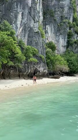 A couple walks on a sandy beach in front of towering limestone cliffs covered in lush green trees. The clear turquoise water gently laps the shore. The man is wearing blue shorts and the woman is wearing a blue bikini. They are walking away from the camera and towards the cliffs.