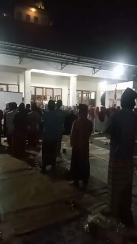 The video shows a group of people standing on mats on the ground at night, facing a building. Many of the individuals are wearing traditional Islamic attire, including peci caps and sarongs. The building appears to be a mosque or a similar religious institution, with a covered walkway and lights illuminating the scene. Some individuals are standing and looking forward, while others appear to be in prayer or in the process of praying. There are also some objects scattered on the ground, possibly bags or personal belongings. The overall atmosphere suggests a religious gathering or ceremony taking place outdoors after dark.