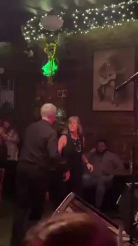 The video shows a couple dancing at a bar. The man and woman are dressed in black and have gray hair. They are dancing close together, and the man is leading. The bar is dimly lit, with string lights and a disco ball hanging from the ceiling. There are other people seated at tables in the background, some watching the couple. A painting of a musician with a guitar is visible on the brick wall. The atmosphere is lively and intimate.