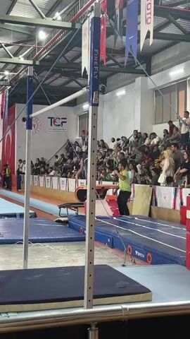 This is a vertical, medium shot video from a gymnastics competition. The focus is on a girl in a blue and yellow leotard performing on a balance beam. She is in the middle of a move and slightly bent over. A coach in a green shirt and black pants stands to her right, looking at her. In the background, spectators are seated in bleachers. The gym environment is visible with equipment like the uneven bars and a vaulting table in the foreground. There are banners and flags hanging from the ceiling. The video cuts off as the gymnast is still in motion.