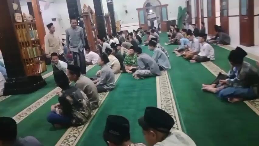 The video shows an interior view of a mosque with a large group of people, mostly young males, sitting on a green carpet. They are arranged in rows, facing forward, with some individuals standing and walking in the aisles. The people are dressed in traditional Islamic attire, including peci caps and long shirts or robes. Some of the individuals appear to be engaged in religious activities, possibly prayer or a lesson, as they are seated in a contemplative manner. The mosque is decorated with patterned carpets and has architectural features typical of Islamic places of worship, such as arches and columns. The lighting suggests it is daytime or a well-lit indoor space. The overall atmosphere appears to be one of community and religious observance.
