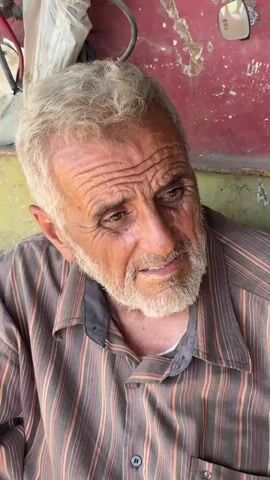 A close-up video shows an elderly man with wrinkled skin and grey hair. He is wearing a striped shirt. His eyes are brown, and he has a grey beard. He appears to be looking slightly to his left. The background is out of focus, but it appears to be a textured wall, possibly red and green, with some white material visible in the upper left corner. The lighting suggests an indoor or shaded outdoor environment.