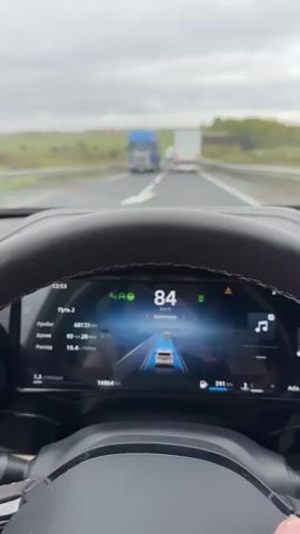 The video shows the view from inside a car, likely the driver's perspective, while driving on a highway. The car is using autopilot, maintaining a speed around 85-91 km/h. In front of the car, there are two large semi-trucks, one blue and one white. The road is a multi-lane highway with a dividing barrier on the left and green fields and trees visible in the background under an overcast sky. The car's dashboard is visible, displaying various driving metrics like speed, fuel consumption, and trip details, presented in Russian.