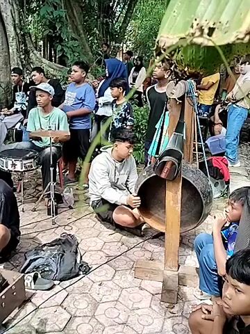 The video shows a group of young men playing traditional Indonesian music outdoors. They are sitting and standing on a patterned stone surface in a park-like setting, with a large banyan tree behind them. The musicians are playing various percussion instruments, including drums, gongs, and a large metal instrument struck with a mallet. Several people are standing around watching and listening to the music. There are various objects scattered around the musicians, including bags, shoes, and musical equipment. The overall atmosphere is relaxed and informal.