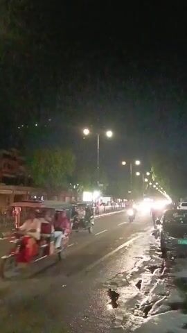 The video depicts a street scene at night, likely after rainfall given the wet pavement. Streetlights illuminate the road, creating reflections on the wet surface. Vehicles, including a tuk-tuk, motorcycles, and cars, are seen moving along the road. Buildings line one side of the street, showing architectural details. Pedestrians are also present.  The overall ambiance suggests an urban setting at night following a recent rain.
