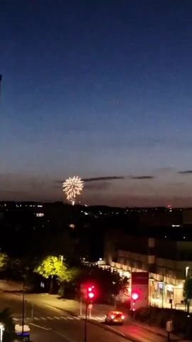 The video shows a fireworks display at dusk or night over a city. The sky is a deep blue, transitioning to a lighter shade near the horizon. In the foreground, a street is visible with traffic lights, one of which is red, and a car driving past. Buildings and trees are silhouetted against the sky. A large firework, with a burst of light and sparks, is visible in the upper left quadrant of the frame. The fireworks are colorful, with yellow and white light emanating from them. The scene appears to be from a high vantage point, possibly a balcony or rooftop.