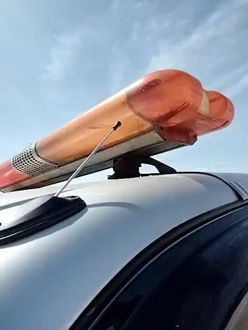The video shows the top of a silver car as it travels on a highway or bridge. The car has an orange emergency light bar on its roof and a small antenna. The sky is blue with some clouds. The car passes by a white tent and some orange traffic barriers on the side of the road. The wind is quite strong. In the distance, a mountain can be seen.