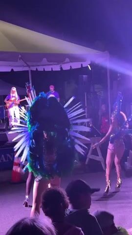 A nighttime outdoor event features a stage with a band and dancers. On the left, a woman with blonde hair plays a guitar. Next to her, a person in a purple shirt appears to be DJing. Center stage, two women in elaborate feathered costumes are dancing. The woman on the left has a large headdress and costume in shades of green and blue, with a sparkly outfit. The woman on the right has a similar headdress and costume in blue and purple, with a gold, bejeweled outfit. They are performing in front of an audience, some of whom are visible in the foreground. The stage is lit with purple lights.