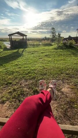 A person wearing red pants and floral slippers sits with their legs crossed on a bench outdoors. In the foreground, a metallic mug of liquid is held by a hand. The background features a grassy area with a wooden gazebo and a body of water. The sun is visible in the sky, casting a warm glow on the scene. The video appears to be a time-lapse or a series of still shots showing the progression of the sun's position in the sky over a short period.