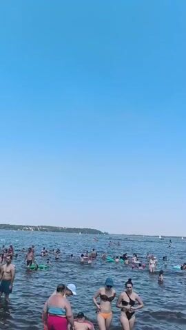 A wide shot of a crowded lake on a clear, sunny day. The sky is a vibrant blue with no clouds. The water is dark and appears to be somewhat choppy. Numerous people are in the water, some wading and others swimming. Many are wearing swimwear. Several individuals are on floatation devices, including inflatable tubes and what appear to be paddleboards. In the background, a treeline is visible on the horizon, and a few sailboats can be seen. In the foreground, three women in bikinis are standing in the water, facing the camera. One woman on the left is wearing a blue hat. Another woman to her right is wearing sunglasses. The woman on the far right is wearing a black bikini. There are other people in the water around them. The overall scene depicts a popular recreational spot on a warm day.