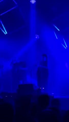 Two musicians perform on a stage illuminated by blue light. A guitarist, seen on the left, is wearing a t-shirt and hat. A second musician stands on the right near a microphone. Behind them are cube-shaped stage lights and spotlights. The backs of heads in the audience are visible in the foreground.