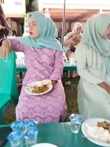 The video shows a gathering of people seated at tables under a canopy, likely at an outdoor event or celebration. The people are dressed in formal attire, with many women wearing hijabs. They appear to be eating and socializing. There are plates of food and drinks on the tables, as well as plastic chairs and red buckets. The environment is outdoors, with visible trees and a building in the background. The lighting suggests it is daytime.