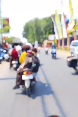 The video shows a blurred street scene with several motorcycles. The motorcycles are moving along a road, and the riders are mostly obscured by the blur. In the background, there are trees and buildings, and some flags are hanging from poles. The lighting suggests it is daytime, and the weather appears to be sunny. The overall impression is a busy street with traffic, possibly in a town or city.