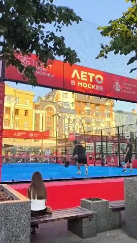 This video shows a padel tennis match taking place outdoors. The court is vibrant blue with red borders, enclosed by glass walls and a net. Two men are actively playing on the court, one in a black t-shirt and shorts, the other in a black t-shirt and darker shorts. Several other people are visible in the background, some spectating from bleachers and others standing near the court.

In the foreground, a woman with long brown hair is seated on a wooden bench, facing away from the camera, observing the game. Two stone planters are situated near the bench.

Above the court, a large red banner stretches across, featuring text in Russian. The prominent phrase "ЛЕТО В МОСКВЕ" (Summer in Moscow) is displayed in large, stylized white lettering, indicating the event's location and season. Smaller text on the banner includes "Департамент спорта города Москвы" (Department of Sports of the City of Moscow) and "МЭРА" (Mayor's).

The setting appears to be urban, with buildings visible behind the court. The lighting suggests it is daytime, likely late afternoon or early evening, as indicated by the warm sunlight casting shadows and illuminating the scene. The presence of trees with green leaves frames the top of the shot, adding a natural element to the urban backdrop. The atmosphere seems relaxed and enjoyable, with people engaged in sport and leisure.