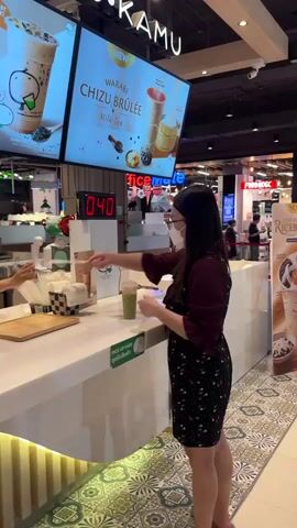 The video shows a woman in a black dress with a floral pattern, wearing glasses and a mask, standing at a counter in what appears to be a food establishment. She is holding a straw and looking at a green beverage in a plastic cup on the counter. Behind her, a large digital display shows the number "040" in red. Another display board above shows images of drinks with the text "WARABI CHIZU BRULEE MILK TEA". To the left of the woman, there is a dispenser of straws and a checkered napkin holder. To the right of the green drink, there is a sign that says "PICK UP HERE". In another segment of the video, a young woman in a green shirt and black apron is seen behind the counter, preparing something in a plastic bag. She is wearing a black cap. The environment appears to be a commercial space with lighting and other displays visible in the background.