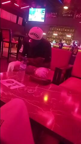 The video shows a person seated at a red, marble-patterned table in a bar or lounge setting. The individual is wearing a dark shirt, white pants, and a white baseball cap with the word "Yankees" printed on it. They appear to be using a mobile phone. On the table, there's a bottle of water, a small bag, and a coaster or button. The lighting in the room is predominantly red, giving the scene a vibrant, almost monochromatic appearance. In the background, there is a bar with a display of bottles and other patrons. A television screen above the bar shows an ongoing broadcast, and there are decorative elements like neon signs and curtains. The general atmosphere seems relaxed and social.