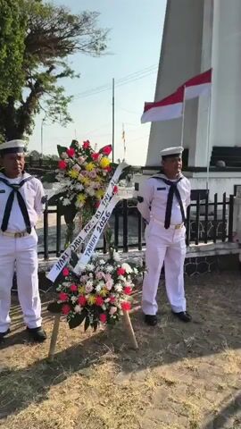 Two men in sailor uniforms stand at attention in front of a floral wreath. The wreath has a white ribbon with text on it. Behind them, a white monument stands with two Indonesian flags flying. The ground is covered in dry grass and dirt. The sky is bright and sunny.