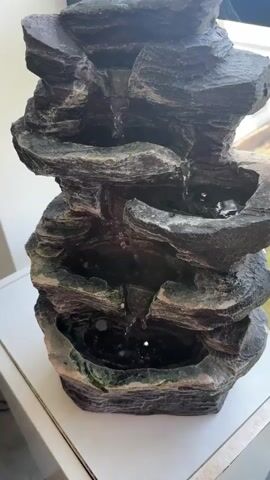 This media file is a close-up, vertical video of a tabletop water fountain designed to mimic a rocky cascade. The fountain is constructed from dark gray material that has been textured and colored to resemble natural stone, with lighter gray and greenish-blue accents suggesting moss or weathering. Water flows from the top of the structure, trickling down multiple tiers and collecting in basins. The movement of the water creates a dynamic visual with reflections and ripples.

The setting appears to be indoors, with a white surface beneath the fountain and a blurred background suggesting a window with natural light coming through. The lighting is soft, indicating it might be daytime, but no specific time of day can be determined with certainty. There are no people or animals visible in the frame. The focus is entirely on the fountain and its operation.

The activity depicted is the continuous flow of water through the fountain, creating a soothing visual and implied soundscape. The notable details include the artificial rock design, the cascading water, and the play of light on the wet surfaces. The overall impression is one of a decorative and possibly calming object.

No visible text is present in the media file. The context provided places the location in Kojetín, Czechia.