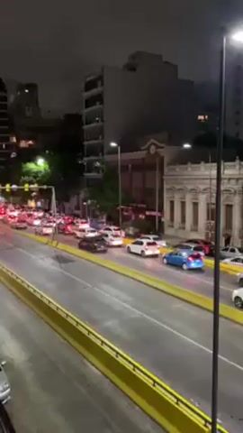 The video shows a street scene at night with heavy traffic. The camera is positioned on an overpass or elevated platform, looking down at a multilane road. The road is filled with cars, mostly white, red, and blue, all stopped at a traffic light. The traffic lights are prominently visible hanging above the road.

The surrounding environment includes tall buildings on either side of the road, with streetlights illuminating the scene. Some trees are also visible, adding a touch of greenery to the urban landscape. The overall ambiance is that of a busy city street during the evening, with the focus on the standstill traffic.
