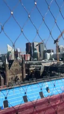 The video shows a view of a city skyline through a metal wire fence. In the background, several modern high-rise buildings are visible, including one with a triangular design. A traditional cathedral with pointed towers is prominently featured below the skyscrapers. In the foreground, scaffolding covered with red and blue tarps suggests ongoing construction or renovation. The sky is clear and blue, indicating a sunny day. The camera focuses on the cityscape, partially obscured by the fence.