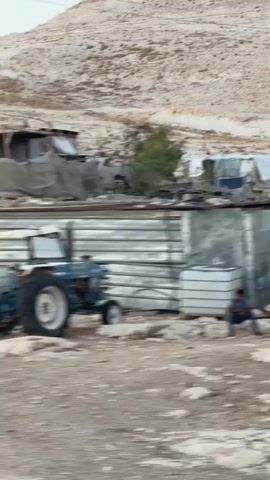The video shows a slow pan of a collection of makeshift structures on a rocky, arid hillside. The structures appear to be constructed from corrugated metal, wood, and other salvaged materials, creating a ramshackle settlement. Some structures have solar panels visible, suggesting an attempt at generating electricity. The ground is covered in loose rocks and debris. The overall impression is one of poverty and rudimentary living conditions. The video appears to be filmed from a moving vehicle, indicated by the motion blur.