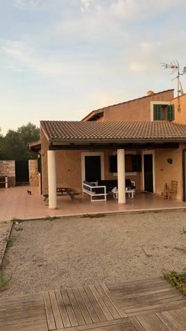 The video pans across the exterior of a peach-colored house with green shutters. A covered patio with white pillars features outdoor furniture. Beyond the patio is a gravel yard with wooden pathways and a variety of plants, including palm trees and succulents. The camera then moves to reveal a swimming pool with turquoise water. A person is swimming in the pool against a backdrop of a sunset with an orange and yellow sky. The environment appears to be rural or semi-rural, with a dry, grassy field and stone walls visible in the distance.