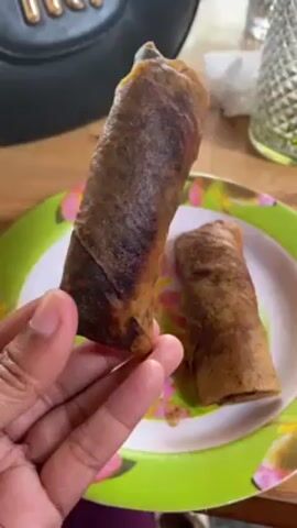 The video shows a hand holding up a fried spring roll. Another spring roll is on a plate with a green, pink, and white floral design. A black handbag sits in the background, along with a glass of water. The spring rolls are dark brown, and the hand is dark-skinned.