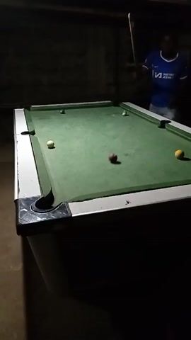 A person wearing a blue shirt and shorts stands on the right side of a rectangular pool table in a dimly lit room. The pool table has a green surface with pockets in each corner and along the sides. There are several pool balls on the table: a white cue ball on the left, a black ball near the top, a red ball and another white ball in the middle, and a yellow ball on the right side. The person is holding a pool cue. The room is dark, and the walls appear to be made of rough material.