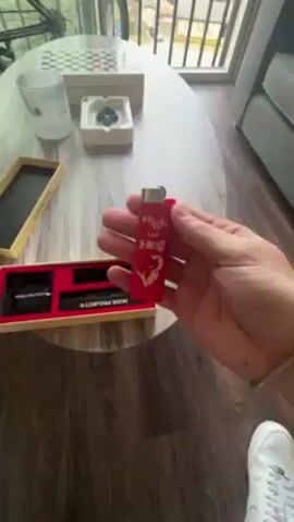 A hand is holding a red lighter with white text that reads "High on Hell" and a devil figure on the side. The lighter has a silver top. The person flicks the lighter and a flame appears. The lighter is held above a box set with a red interior, which contains a roll of protection film and a black cylindrical object labeled "NUDE PROJECT". On the table nearby is an ashtray with a spade design, a glass, a box, and a chess board. A sliver of a gray couch is visible in the background. The floor is wood.