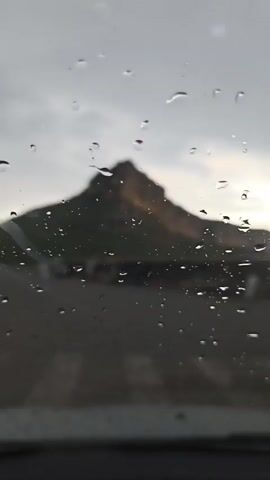 A video shot through a window shows raindrops on the glass, obscuring a distant mountain. The mountain has a distinctive peaked shape, and parts of it are illuminated by sunlight. The foreground is dark, suggesting the view is from inside a vehicle. The sky appears overcast.