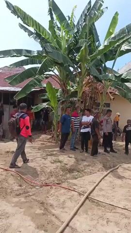 A group of people are gathered outdoors in what appears to be a residential area. There are several houses visible, some with red tiled roofs. Lush green banana trees are prominent in the foreground. The ground is covered in dry, sandy soil with some scattered hoses and buckets. Several men, some wearing casual clothing and others in what looks like a police uniform, are standing around and observing. One man is holding a hose and spraying water onto a structure that appears to be a shed or an extension of a building, which shows signs of fire damage, including charred wood and blackened walls. Another person is visible in the background of the damaged structure, also appearing to be involved in some activity. The sky is clear and blue, indicating daylight. The overall scene suggests an aftermath of a fire, with efforts to extinguish or cool down the damaged area.