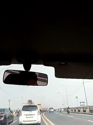 The video is a first-person perspective from inside a car, looking forward and slightly upwards through the windshield. A rearview mirror is visible in the center of the frame, reflecting the face of the driver or passenger. The car is on a multi-lane highway, with other vehicles, including cars, a white van with a round object on its roof, and motorcycles, visible in front. The road appears to be an elevated highway, with concrete barriers and streetlights lining the sides. The sky is overcast and bright, suggesting daytime.