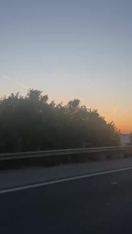 The video shows a sunrise viewed from a moving vehicle. The sky is a gradient of colors from light blue to orange near the horizon, with a few contrails visible. Trees are silhouetted against the bright sun, which is partially obscured by the foliage. A metal guardrail is along the side of the road. The road surface is visible in the foreground. The sound of wind is prominent in the audio.