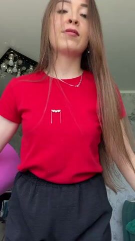 A young woman with long brown hair, wearing a red t-shirt and black pants, stands in front of a light-colored wall. She holds a large bouquet of pink flowers, which she rotates and presents to the camera. The bouquet is wrapped in bright pink paper. In the background, a ceiling light fixture with multiple bulbs is visible, along with a glimpse of pink and green furniture. The woman's expression is neutral as she showcases the flowers.