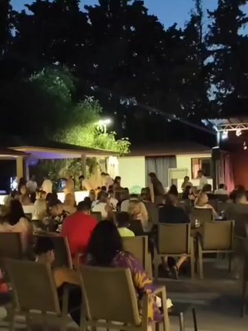 The video shows an outdoor gathering at dusk. A crowd of people are seated at tables, facing a stage area. Lights are set up on a truss above the stage. The sky is dark blue, indicating it is evening. Trees are visible in the background. Some people are standing near the stage. The overall atmosphere appears to be that of an event or performance.