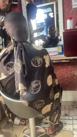 The video shows a barber shop. In the foreground, a young Black person is sitting in a barber chair, facing away from the camera and towards a mirror. They are wearing a dark cape with a light-colored pattern of cartoon bears and possibly other objects. The barber, also a young Black person, is standing behind them, reflected in the mirror, and appears to be cutting their hair with scissors. Another person's face is partially visible in the upper left corner, looking down at the person in the chair. The background of the shop includes a mirror, shelves with various items like bottles and cans, and a red-colored wall. The floor is tiled. The overall scene depicts a typical haircutting service in a barbershop.