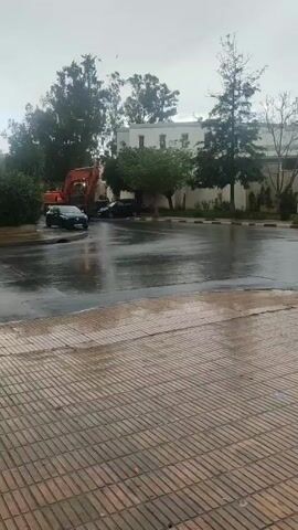 The video shows a rainy scene with a large orange excavator stopped on a wet asphalt road. A black car is parked nearby. In the background, there are trees and a white building. A person in a gray robe walks along the side of the road. The foreground is a paved area with a brick pattern.
